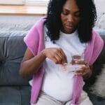 Ciężarna kobieta na sofie popija wodę i przyjmuje witaminy prenatalne